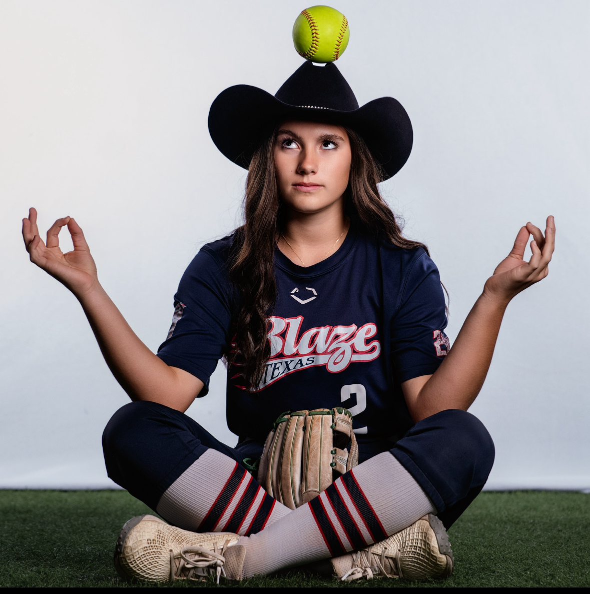 Kaylee Covanes sitting cross-legged in her Blaze Texas jersey balancing a softball on a cowboy hat