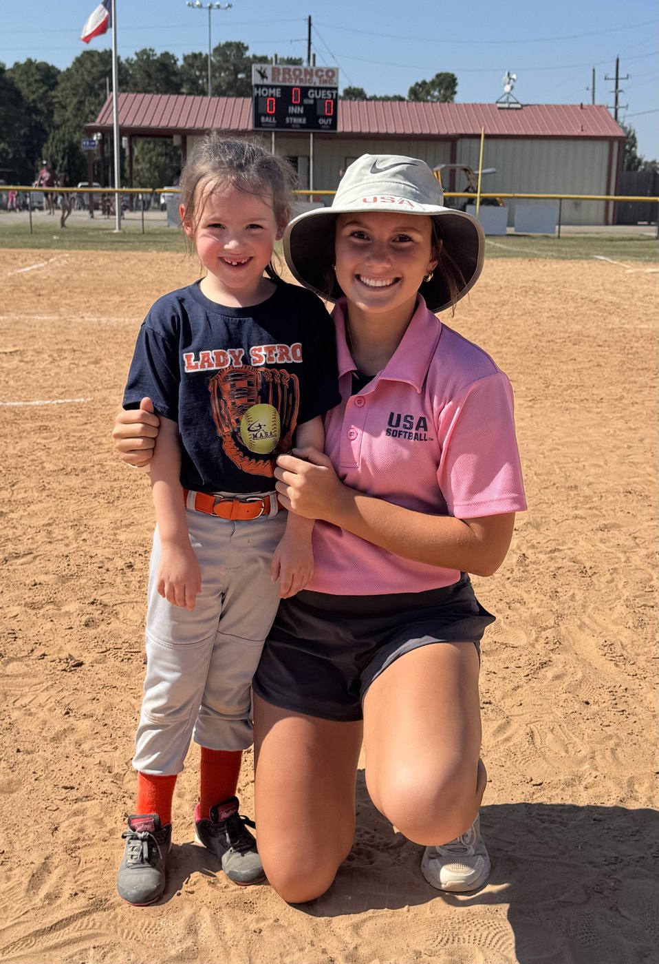 Kaylee Covanes in her USA Softball umpire gear kneeling beside a young player on the infield